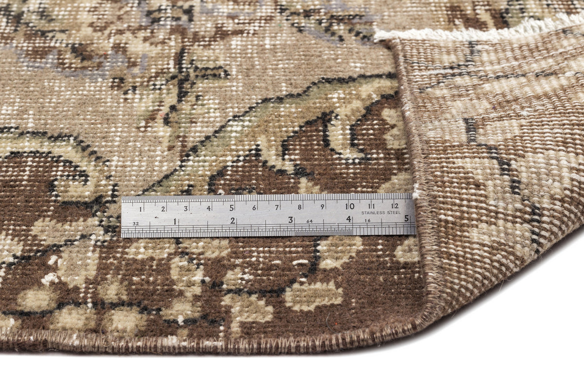Brown and beige vintage-style rug with a distressed floral pattern, shown with a metal ruler for scale.