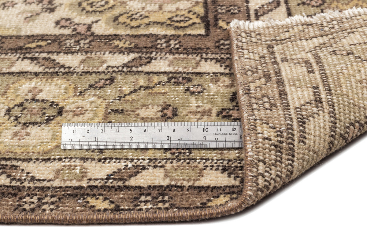 Beige and brown vintage-style rug with a floral pattern, showing a corner fold and a metal ruler for scale.