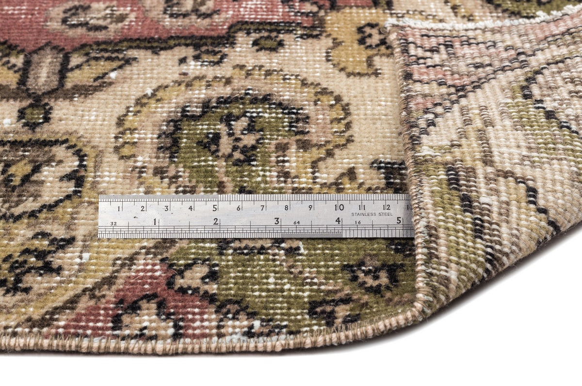 Distressed vintage-style rug with a floral medallion pattern in olive, beige, and muted red, shown with a metal ruler.
