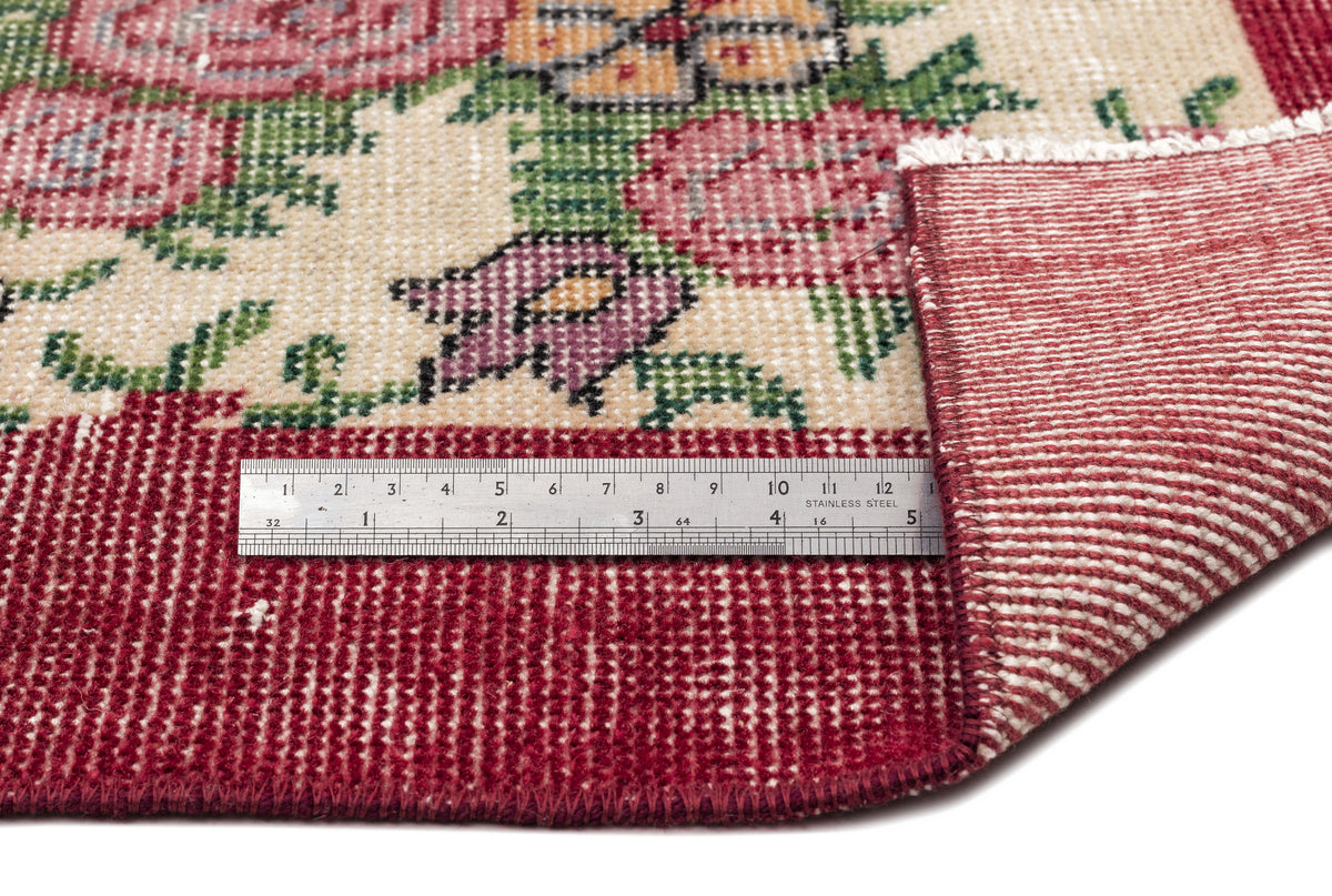 Cream rug with floral pattern in red, green, and purple, featuring a red border and a metal ruler for scale.