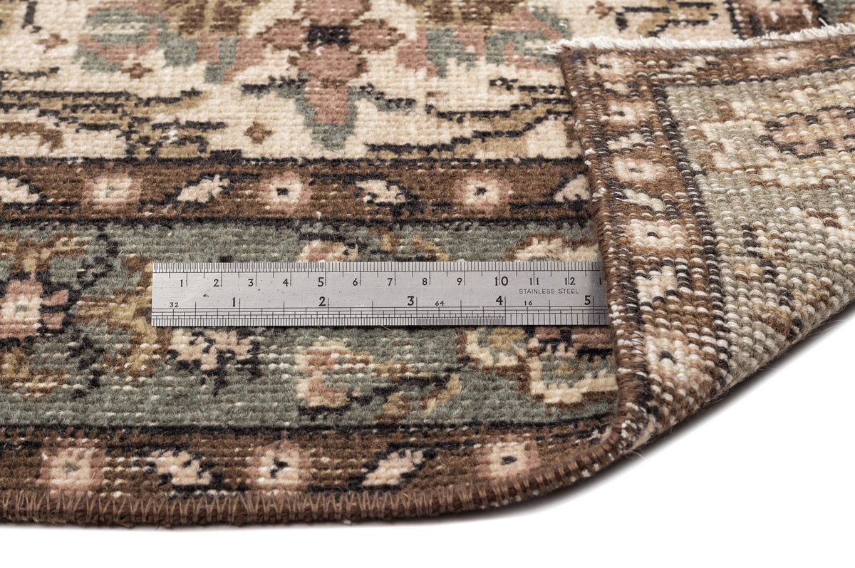 Beige, sage green, and brown traditional patterned rug with a folded corner and a metal ruler for scale.