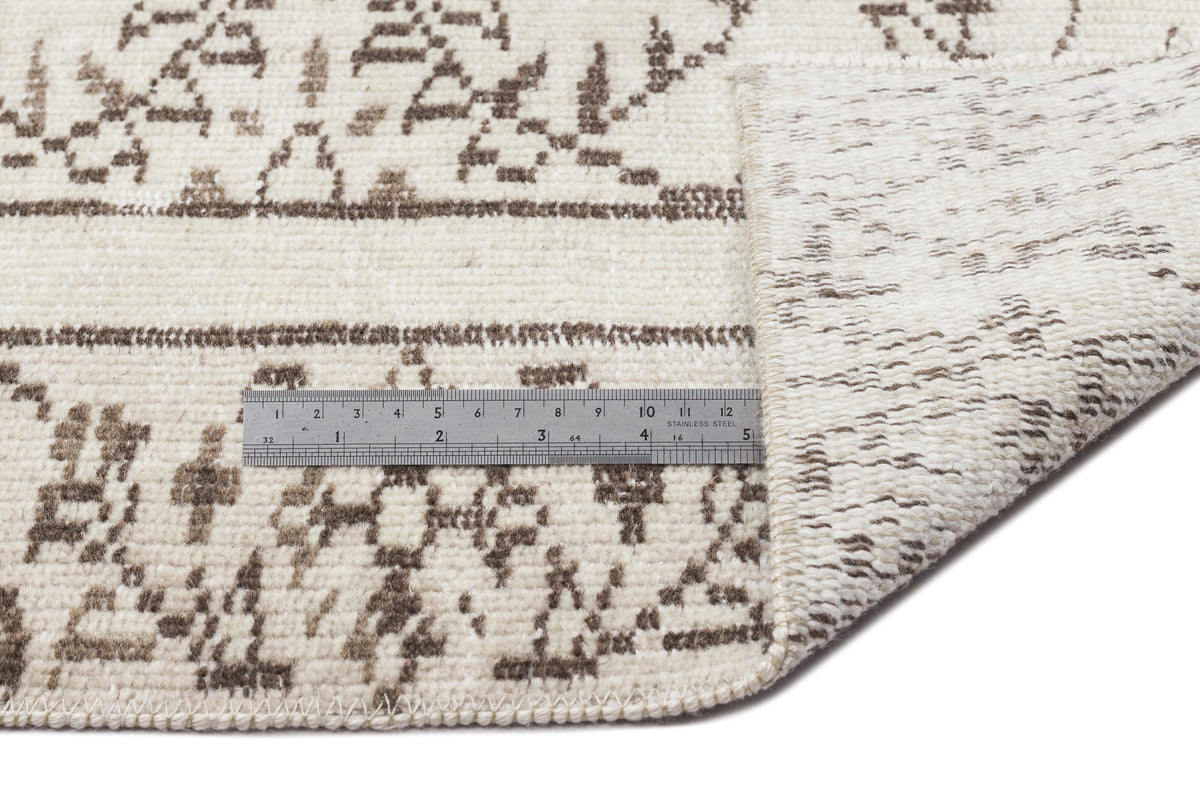 A metal ruler rests on the Greunoa - Vintage Floral Rug - 5'4 X 9'6, which features a folded corner to highlight its thickness and detailed cream and brown patterned texture.