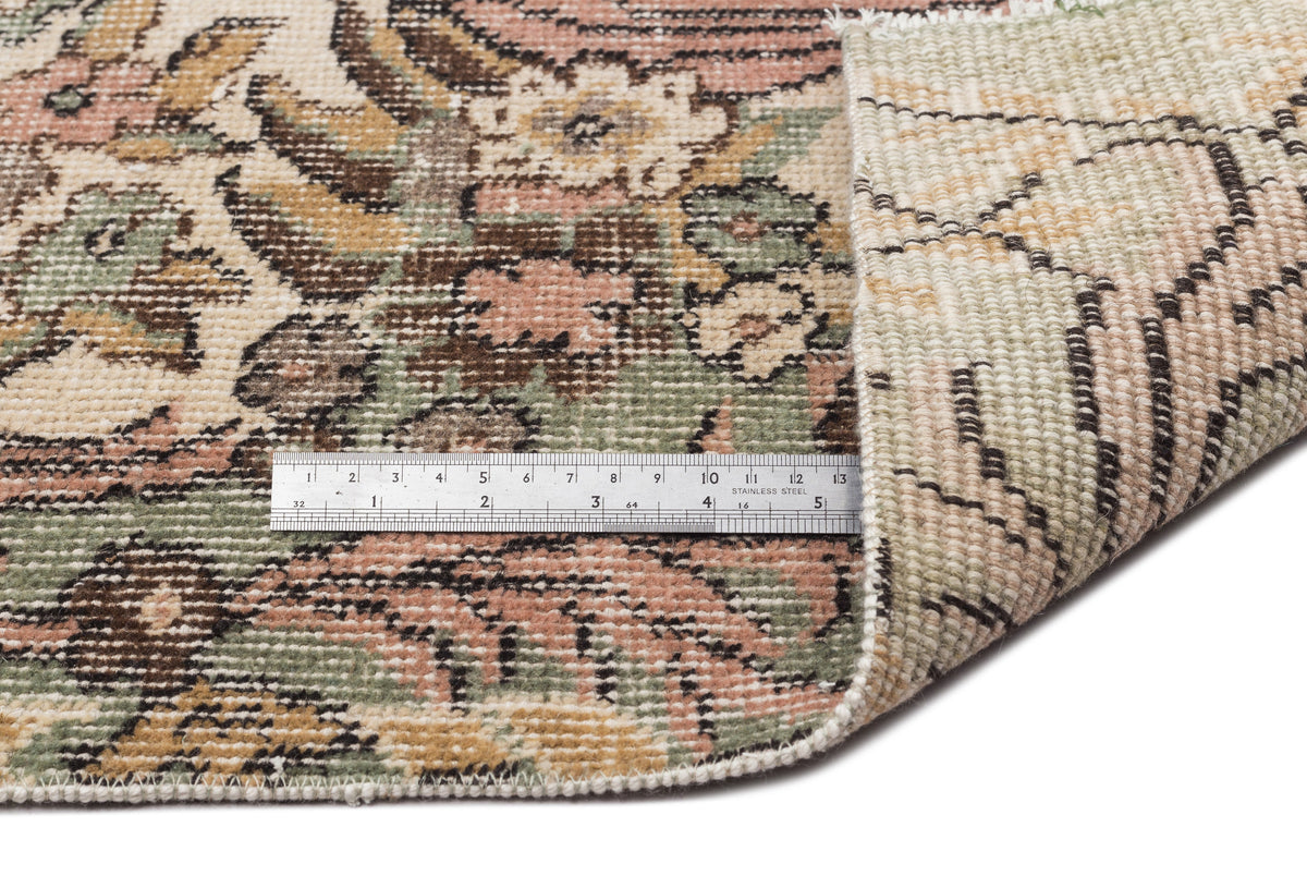 Vintage-style floral rug in muted pink, green, and beige with a metal ruler showing pile texture and backing.