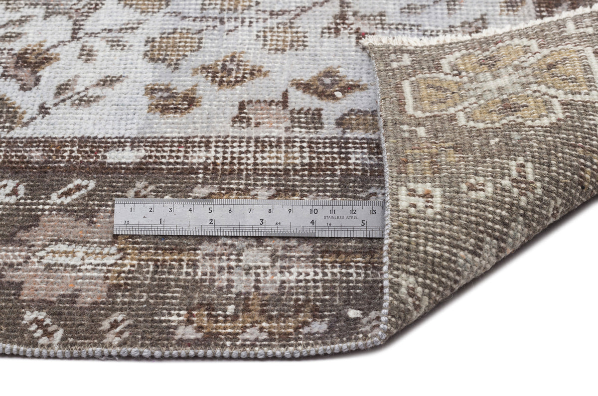 Brown and grey distressed floral rug with a folded corner and a metal ruler for scale on a white background.