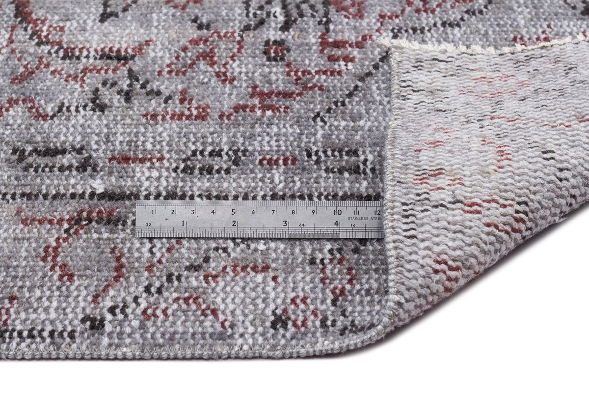 Grey, red, and black distressed floral rug with a folded corner showing the backing and a metal ruler for scale.