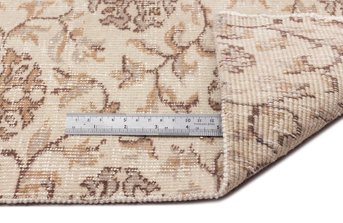 Beige and brown floral patterned rug with a folded corner and a metal ruler for scale on a white background.