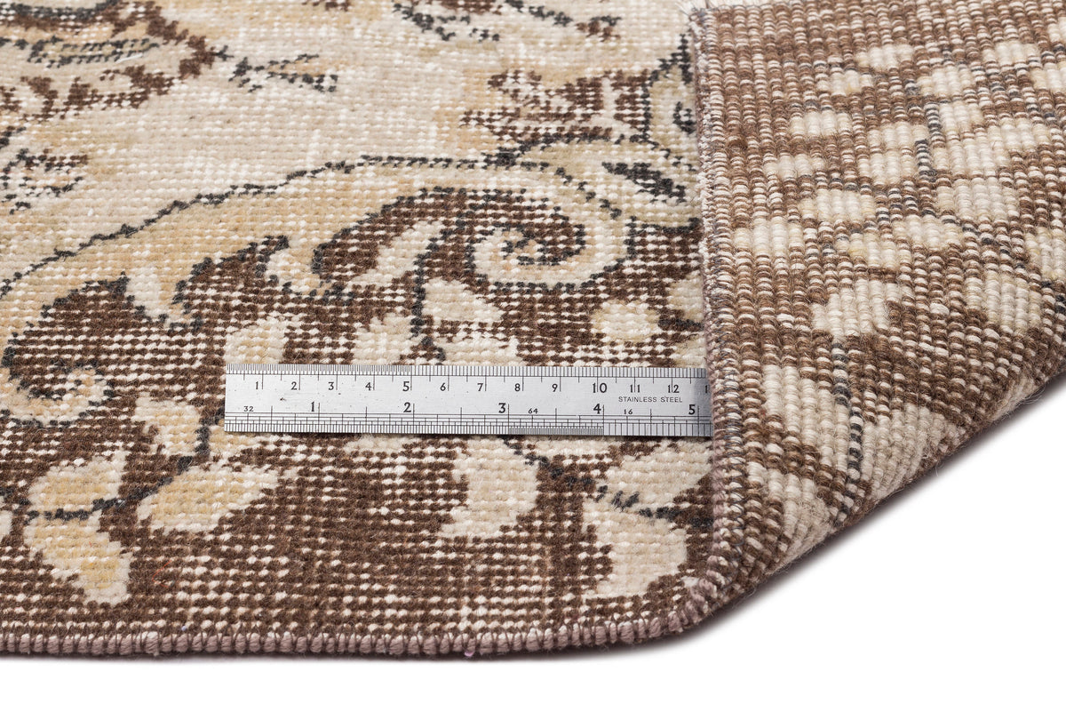 A close-up of the Petida - Vintage Medallion Rug (5'8 x 9'5) shows beige, brown, and cream floral designs with a folded corner revealing the underside. A metal ruler measures in centimeters and inches for scale.