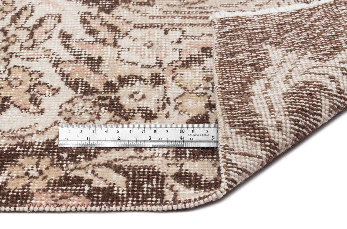 Brown and beige distressed floral rug with a folded corner and a metal ruler for scale on a white background.