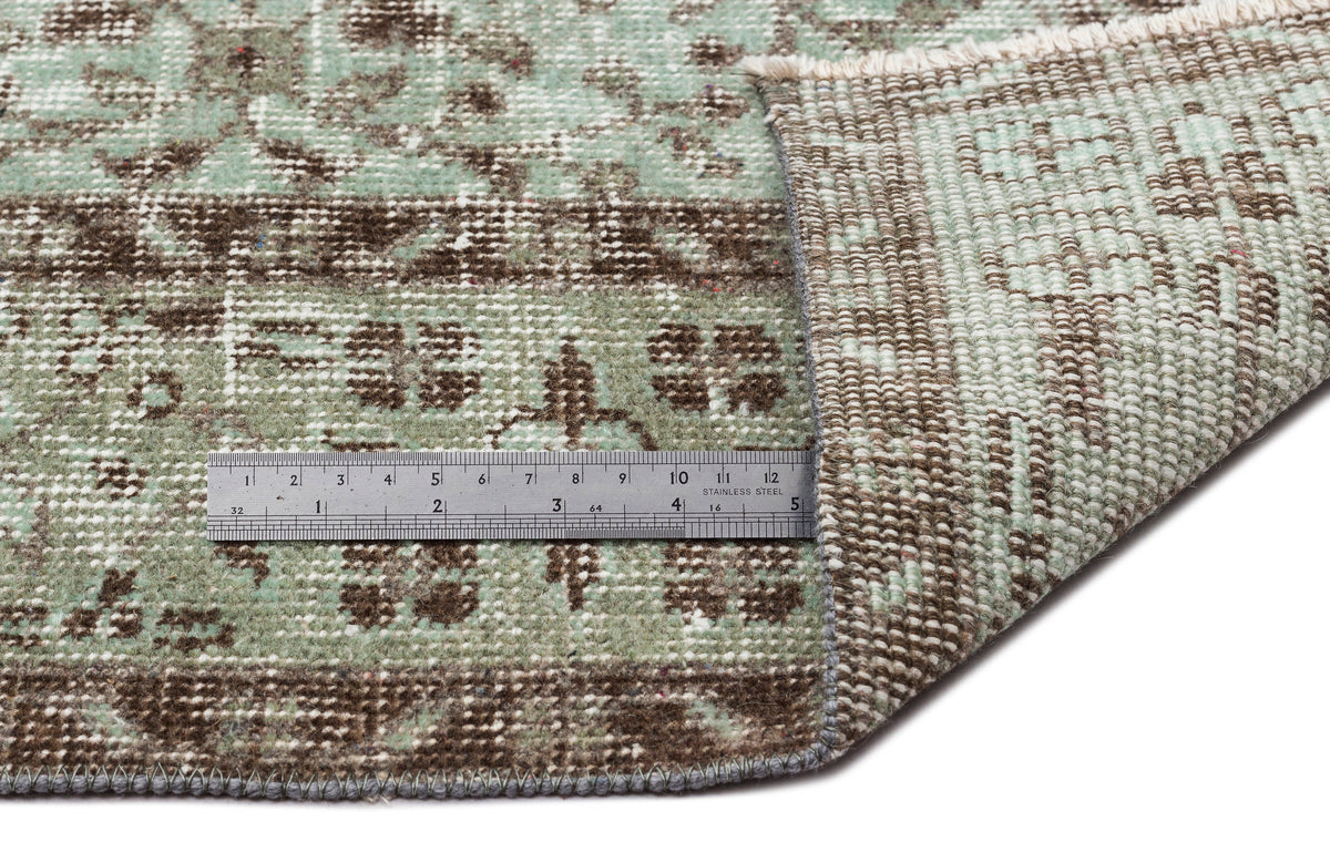 Sage green and brown distressed traditional rug with a folded corner and a metal ruler for scale.