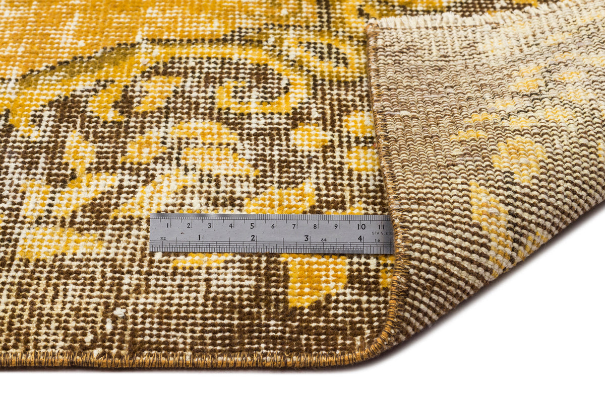 Mustard yellow and brown distressed floral rug with a folded corner and a metal ruler for scale on a white background.