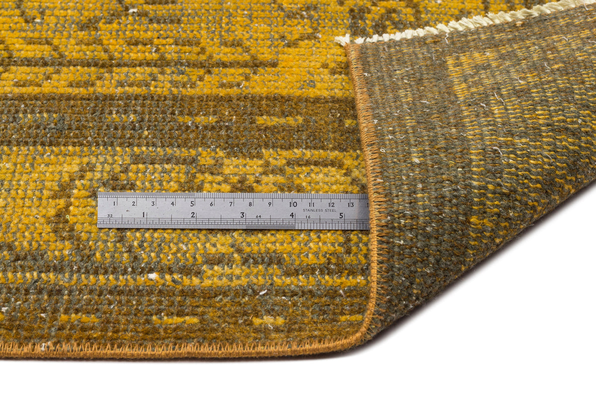Mustard yellow and olive green distressed patterned rug with a folded corner and a metal ruler for scale.