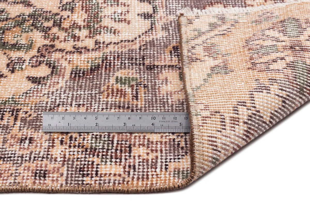 A close-up of the Detiar Vintage Medallion Rug (5'9 X 9'5) corner folded to reveal its underside. A metal ruler beside it shows inches and centimeters for scale. The rug displays a mix of brown, beige, and green tones.
