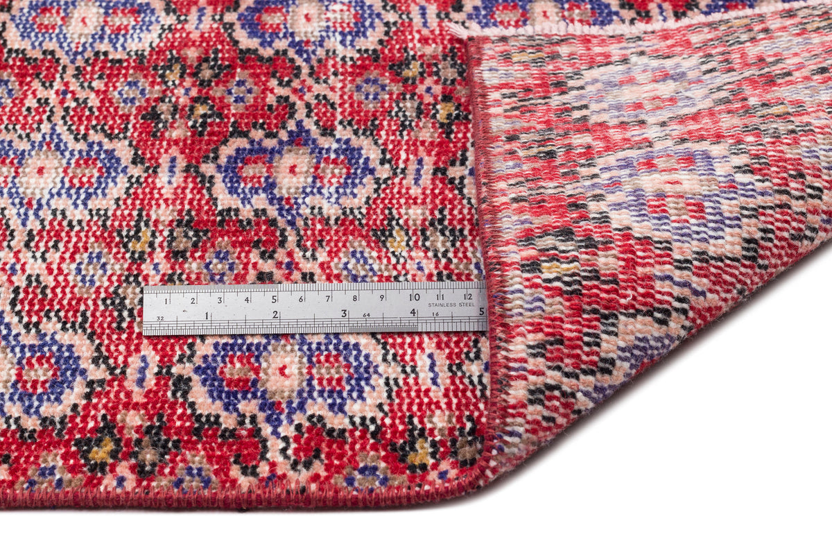 Red, blue, and black traditional floral medallion rug with a folded corner and a metal ruler for scale.
