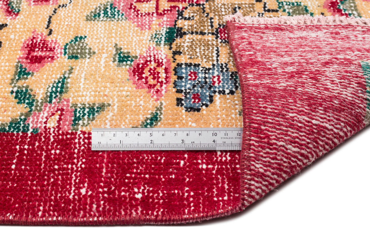 Floral patterned rug with a yellow field and red border, shown folded to reveal the underside with a metal ruler.
