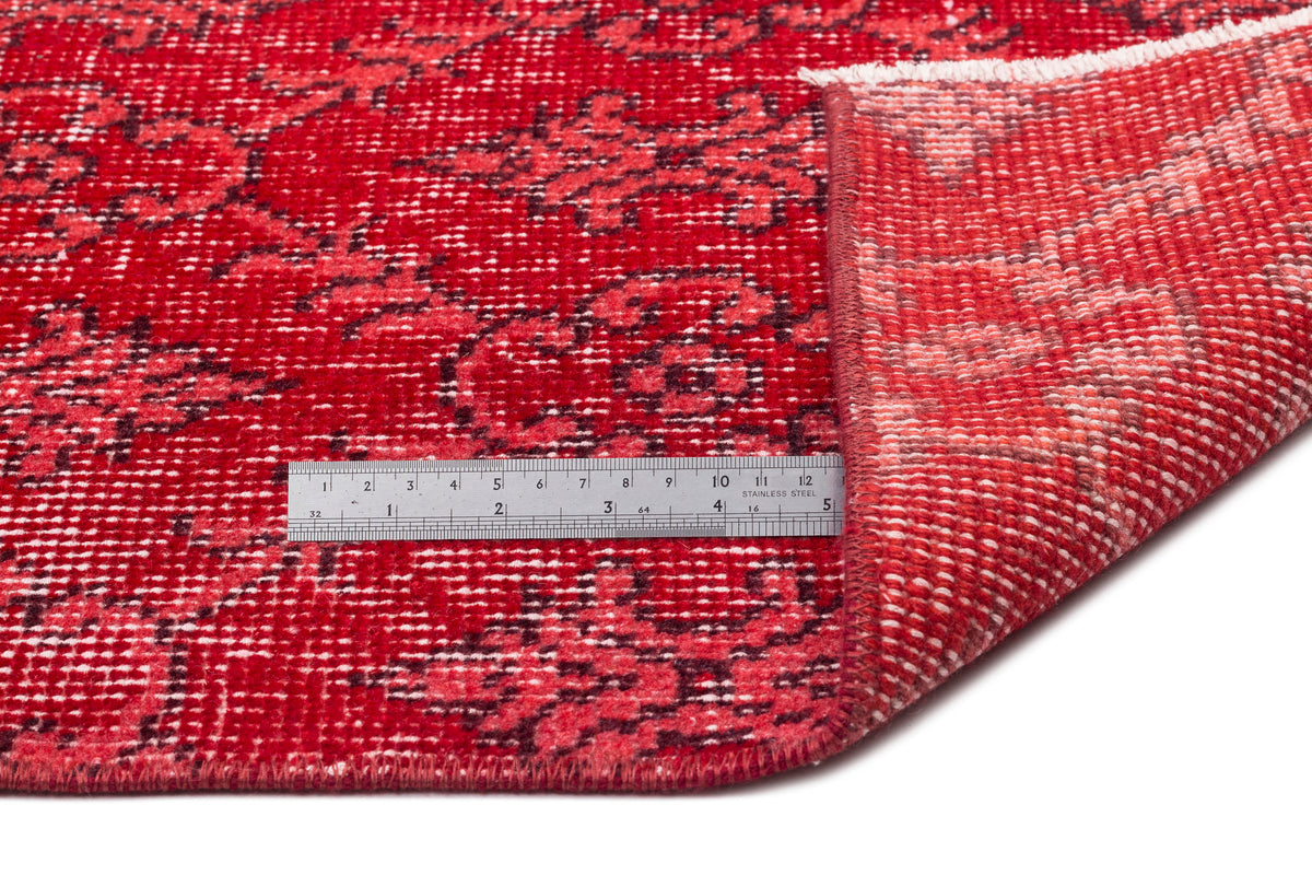 Red and black distressed floral medallion rug with a folded corner and a metal ruler for scale on a white background.