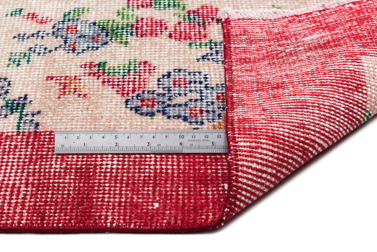 Beige rug with a floral pattern in red, blue, and green, showing a folded corner and a metal ruler for scale.