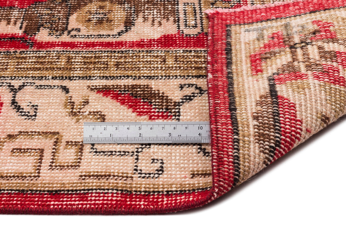 Red, beige, and brown hand-knotted rug with a traditional geometric border, shown with a metal ruler for scale.