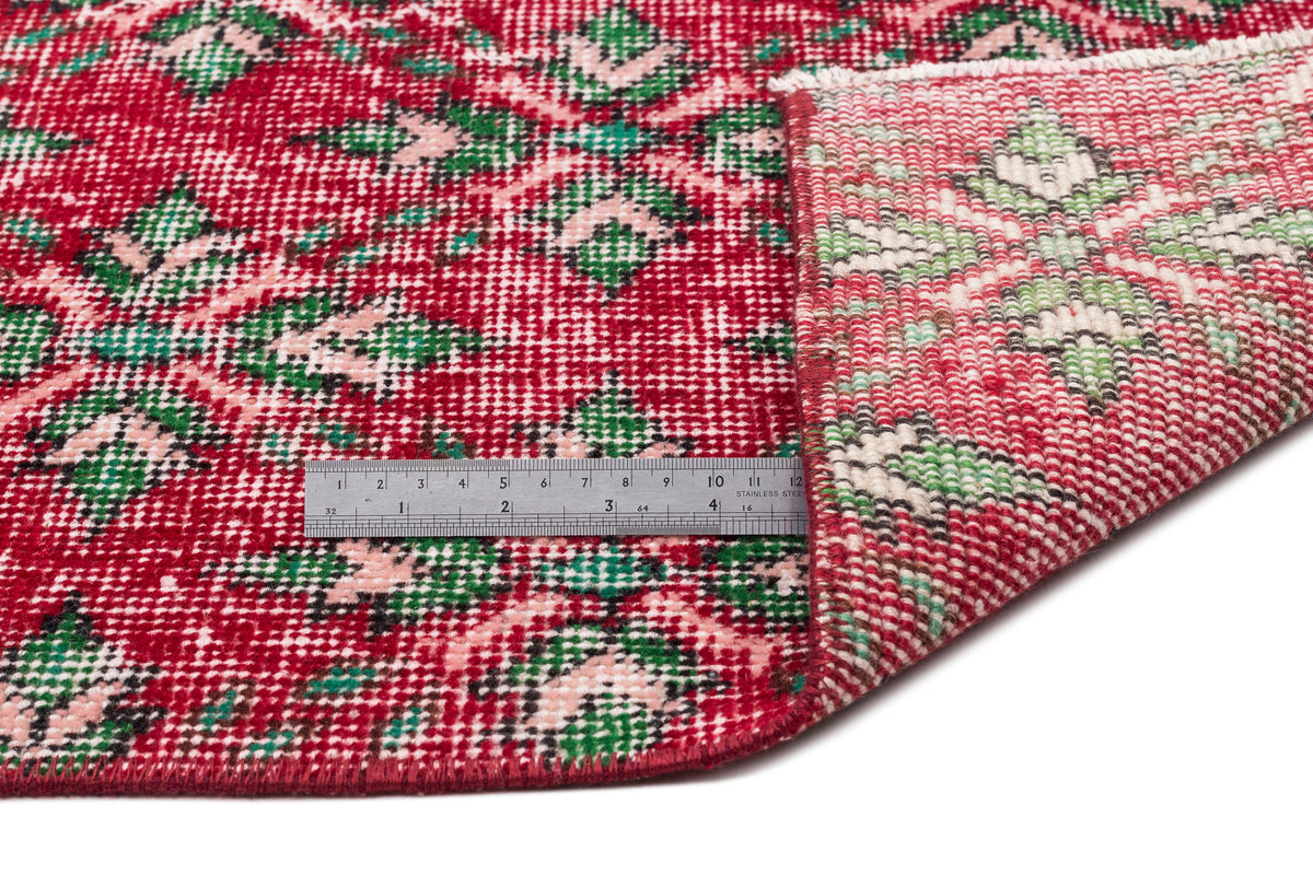 Red, green, and cream floral medallion rug with a folded corner and a metal ruler for scale on a white background.