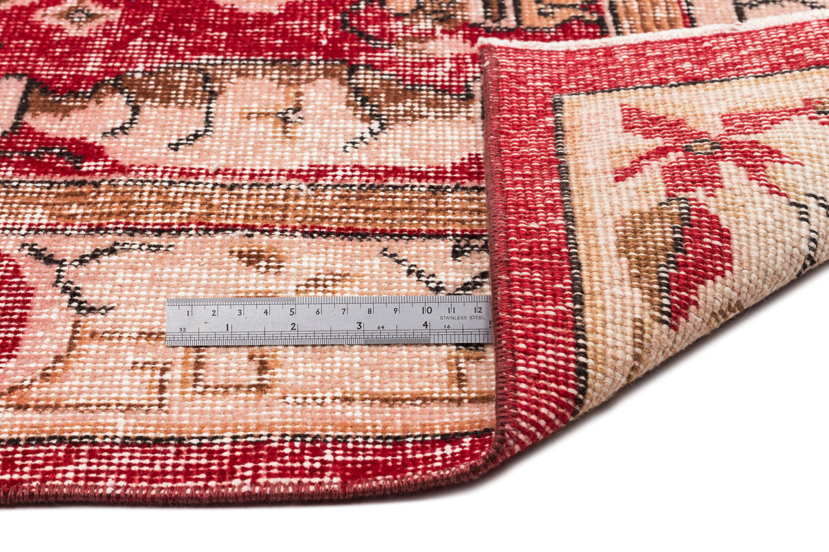 Red and beige traditional rug with a folded corner and a metal ruler for scale on a white background.