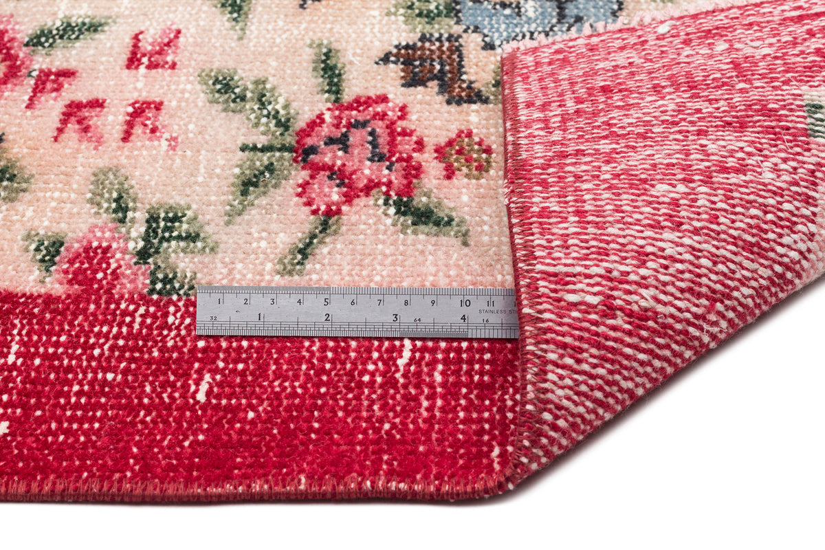 Pink and red floral patterned rug with a folded corner revealing the woven backing and a metal ruler for scale.