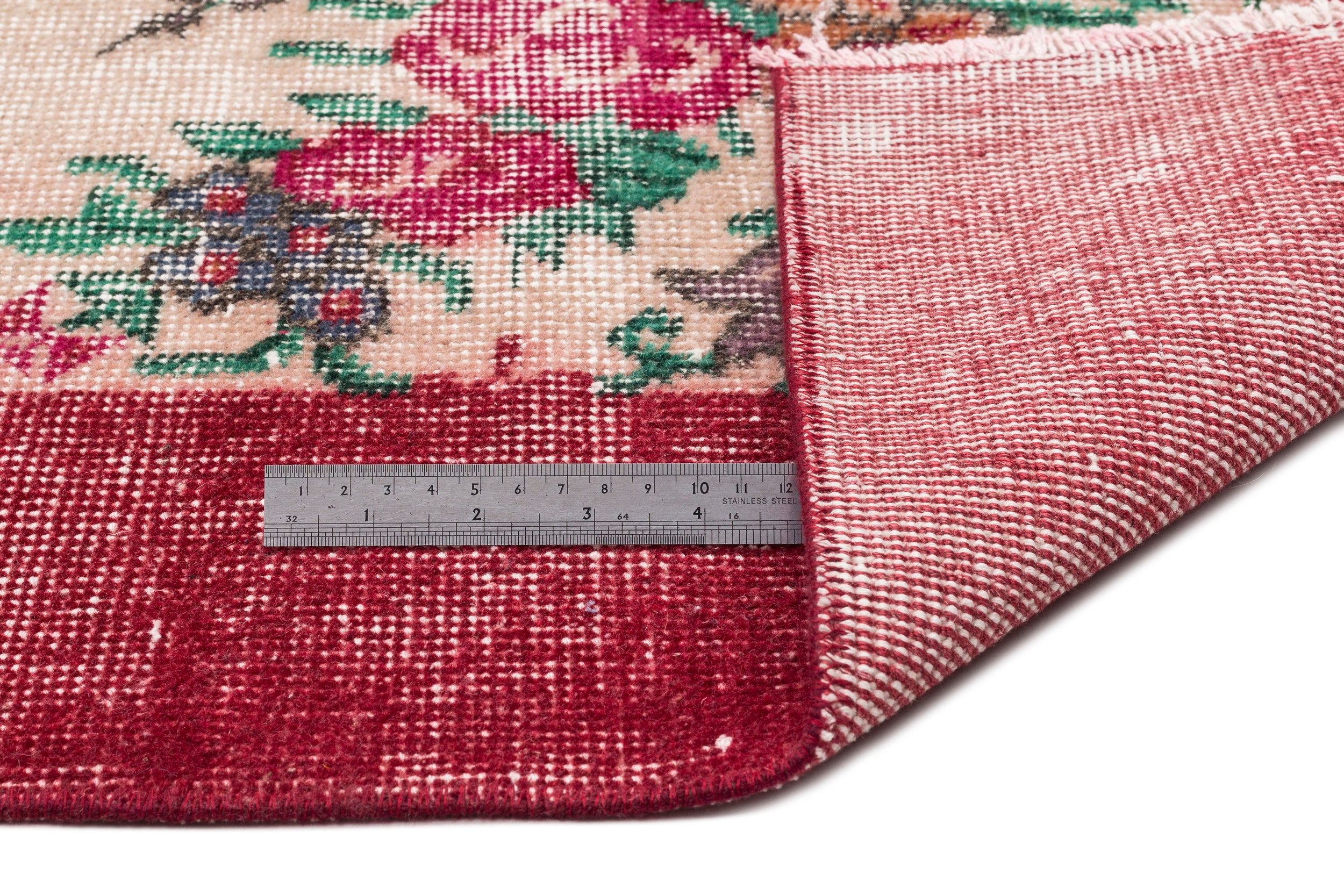 Floral patterned rug with red, green, and cream tones, showing the underside and a metal ruler for scale.