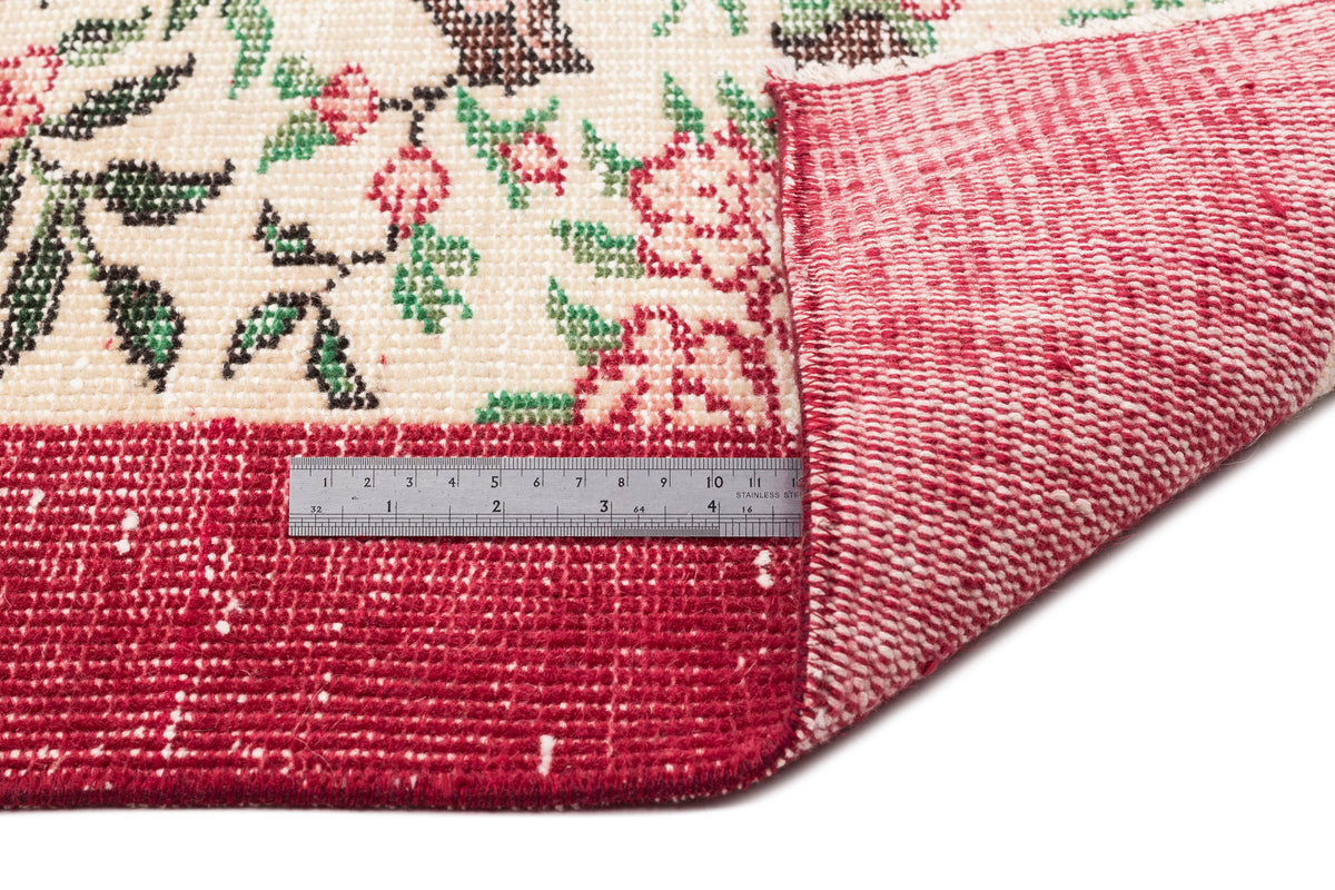 Cream rug with red, green, and brown floral pattern, folded to show the red woven backing and a metal ruler for scale.