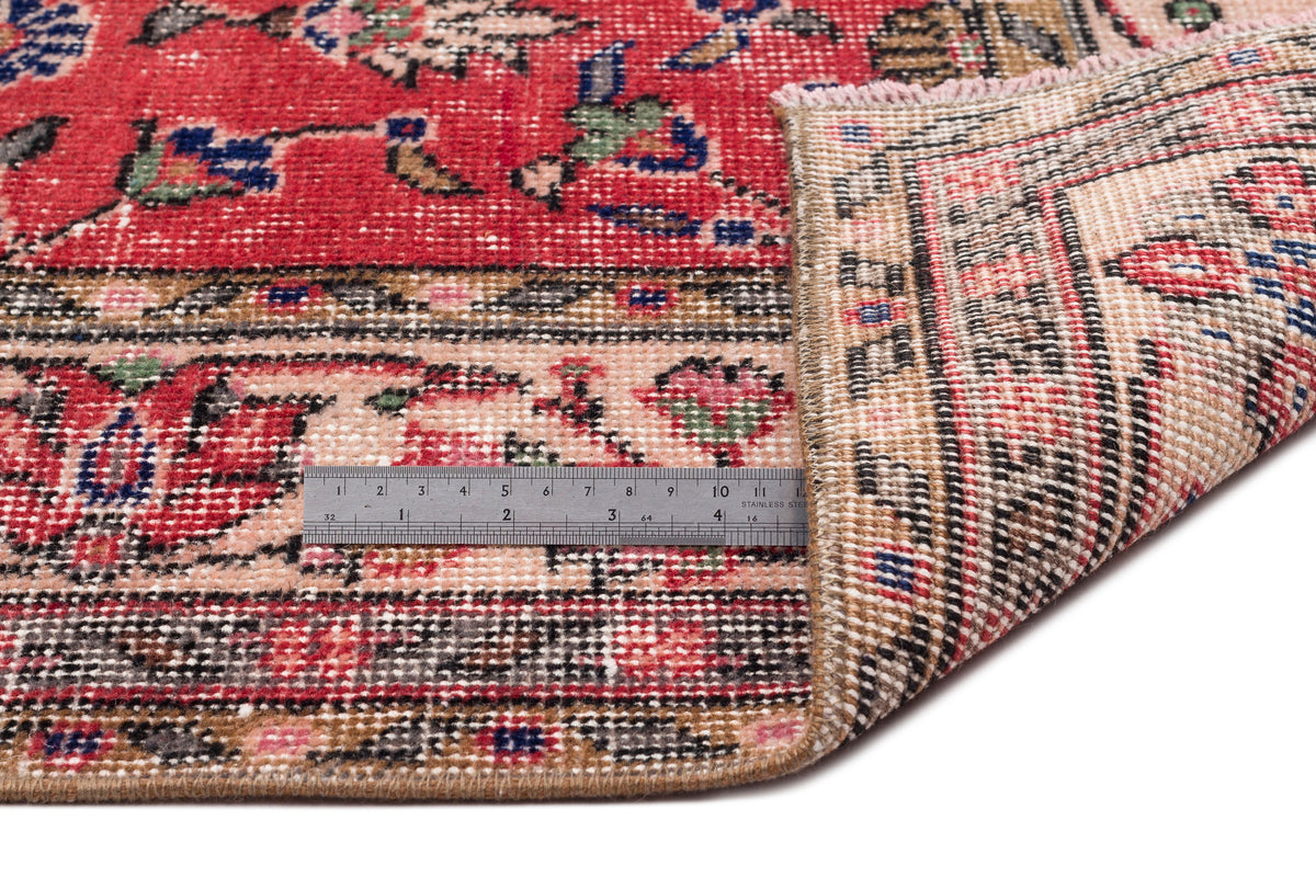 Red and beige traditional rug with floral motifs, shown folded to reveal the weave and a metal ruler for scale.