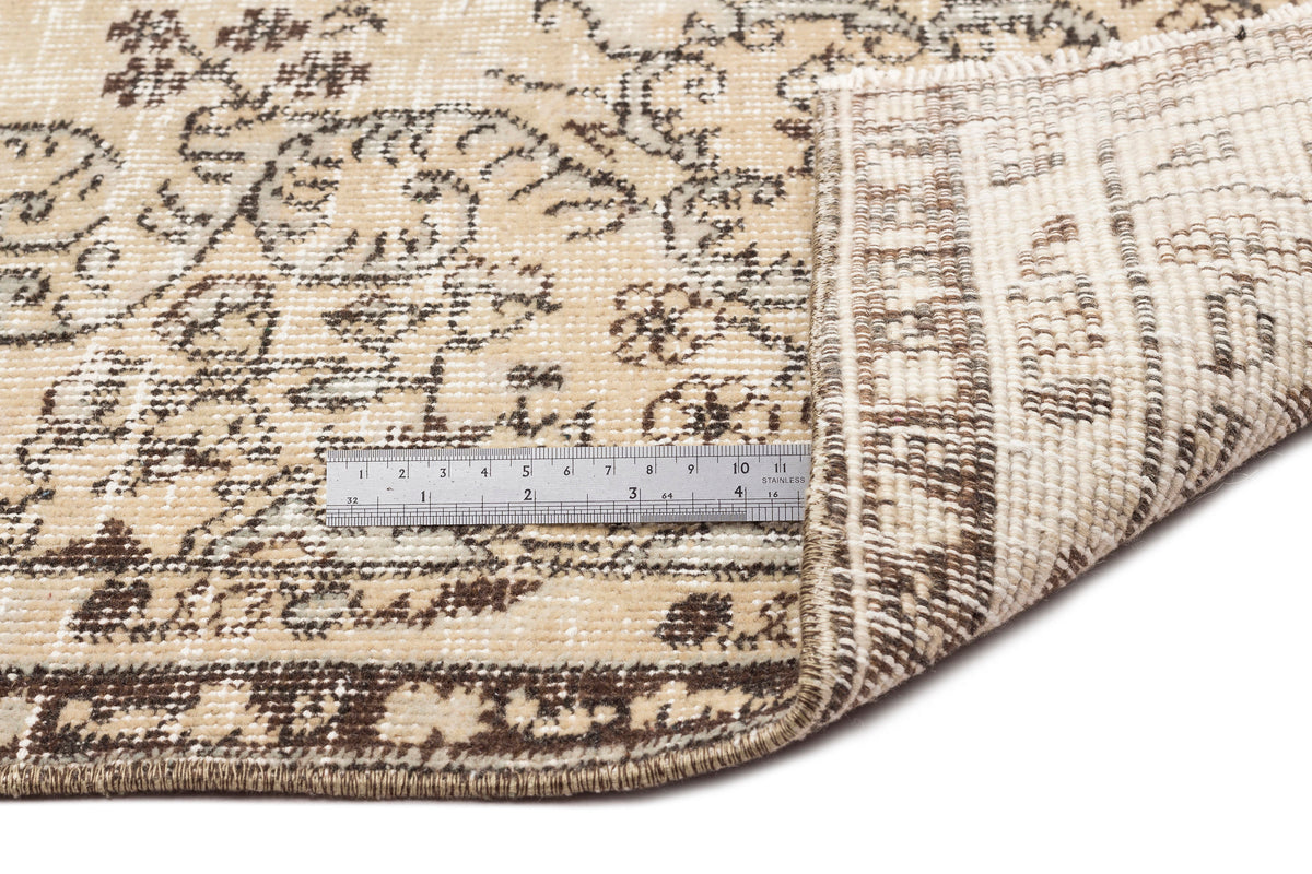 A close-up of the Preutata - Vintage Medallion Rug (5'4 x 8'11) shows its beige and brown pattern, one corner folded, and a metal ruler on top indicating a length of about 15 centimeters for scale.