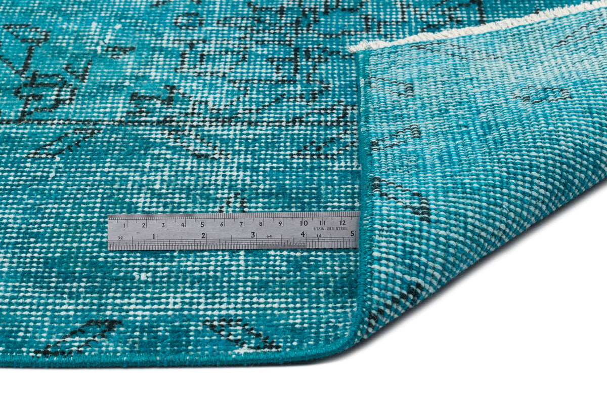 Teal and black distressed patterned rug with a folded corner and a metal ruler for scale on a white background.