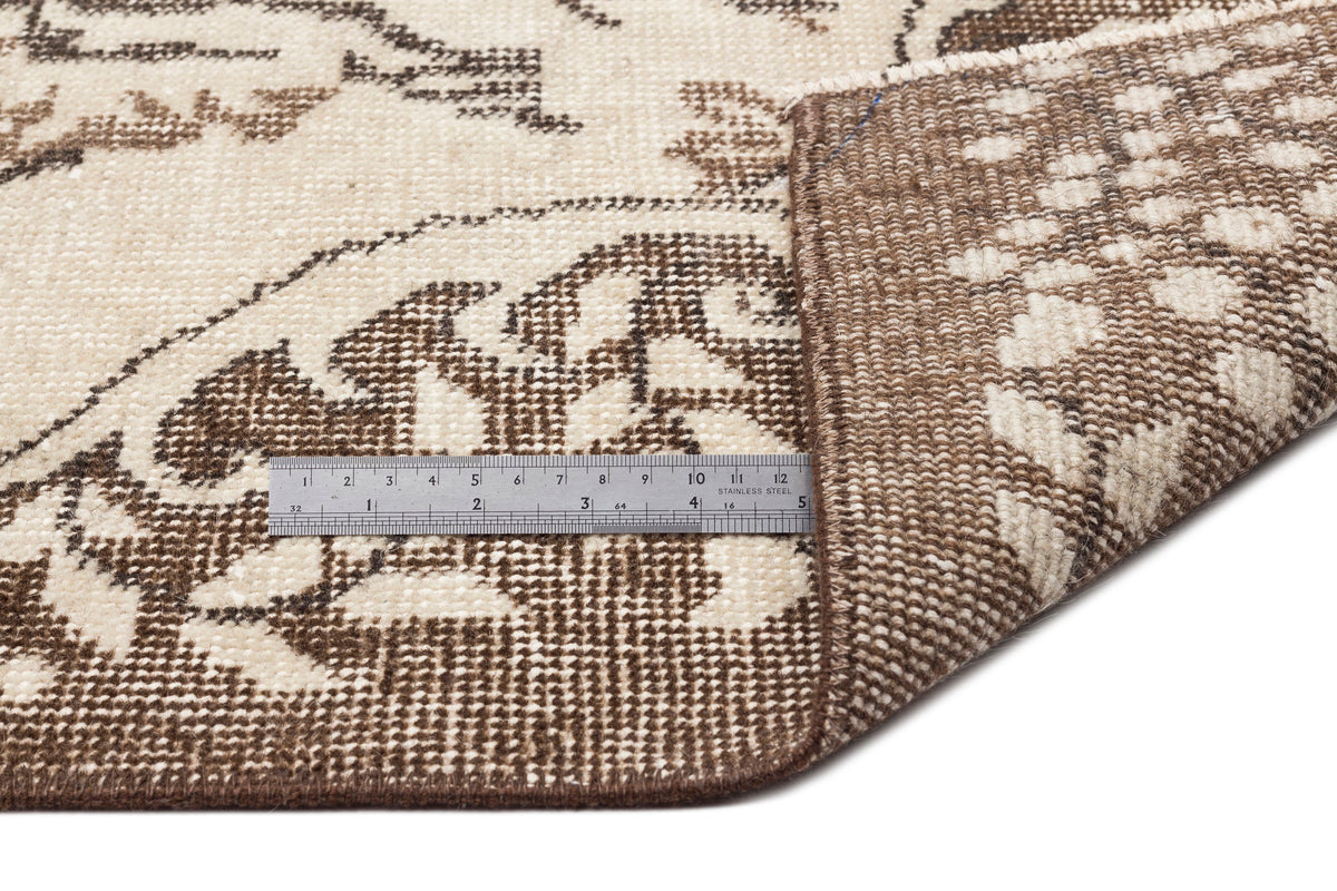 A metallic ruler measures the thickness of the Tiduex - Vintage Medallion Rug - 6'3 X 9'11 near its folded corner, highlighting its beige and brown patterned design with clear ruler markings.