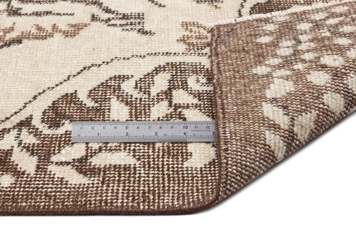 Load image 1 in gallery view. A metallic ruler measures the thickness of the Tiduex - Vintage Medallion Rug - 6'3 X 9'11 near its folded corner, highlighting its beige and brown patterned design with clear ruler markings.