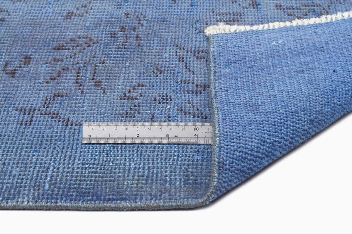 Blue distressed rug with a dark grey floral pattern, showing the textured weave and underside with a metal ruler.