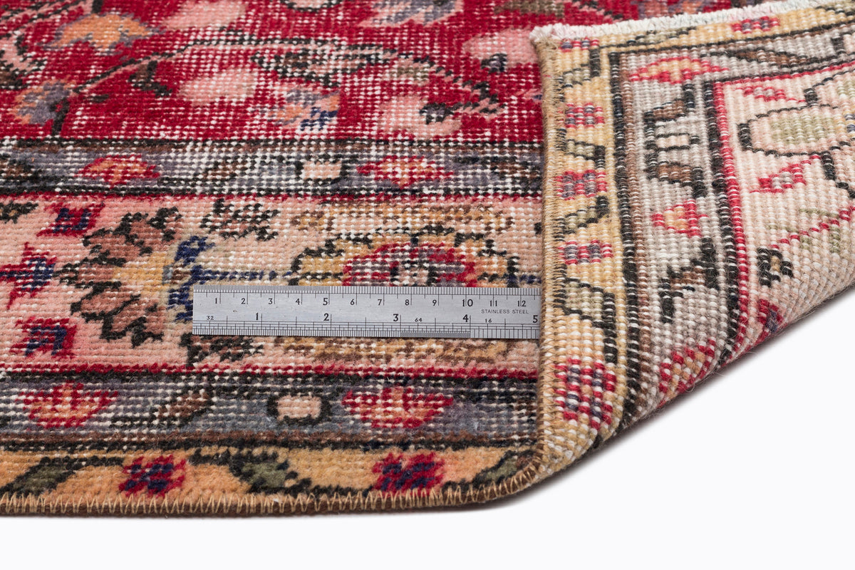 Red and beige vintage-style rug with a floral medallion pattern, shown folded to reveal the underside and a metal ruler.