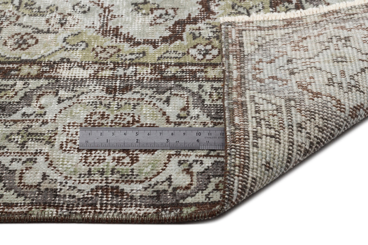 Distressed sage green and brown traditional medallion rug with a corner folded back to show the woven backing and ruler.