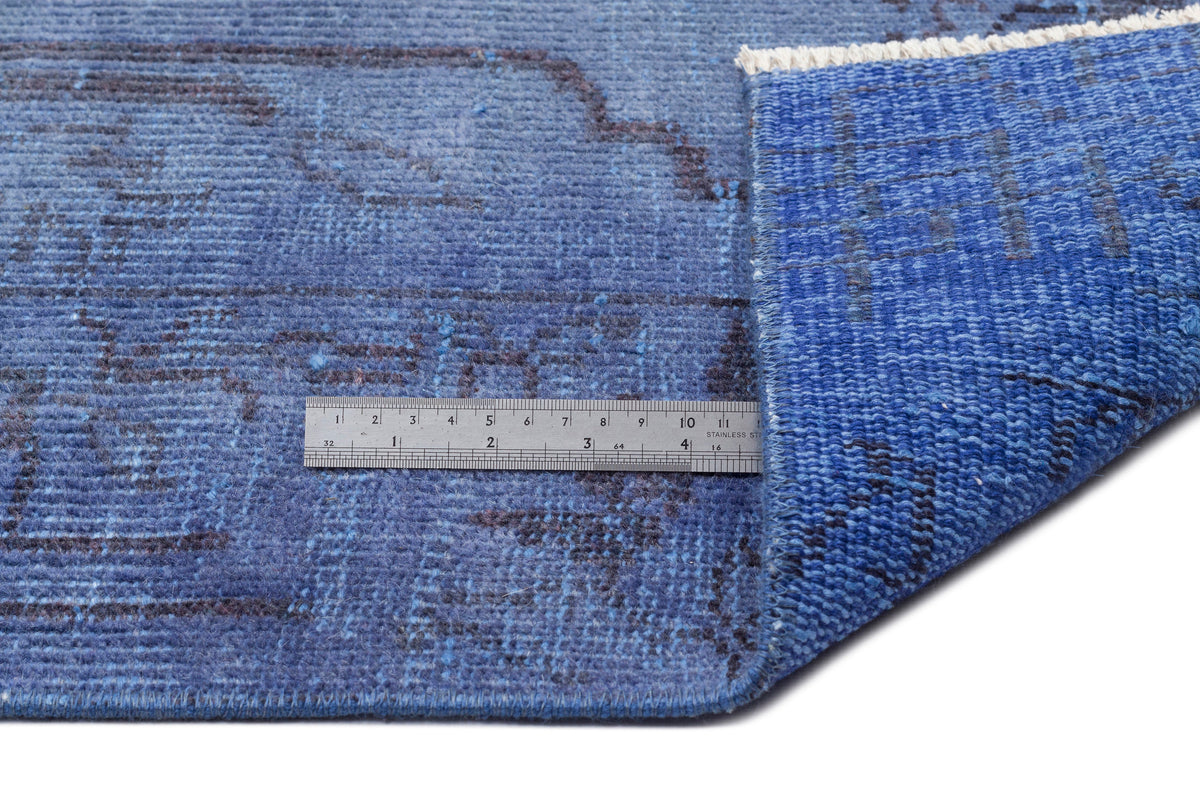 Blue rug with a dark charcoal distressed pattern, shown folded with a metal ruler for scale on a white background.