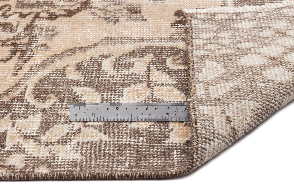 A close-up of the Hakeon - Vintage Medallion Rug (6'5 x 9'8) corner lifted to reveal the underside, with a metal ruler on top for scale. The rug displays beige and brown tones in a floral, geometric, and medallion motif design.