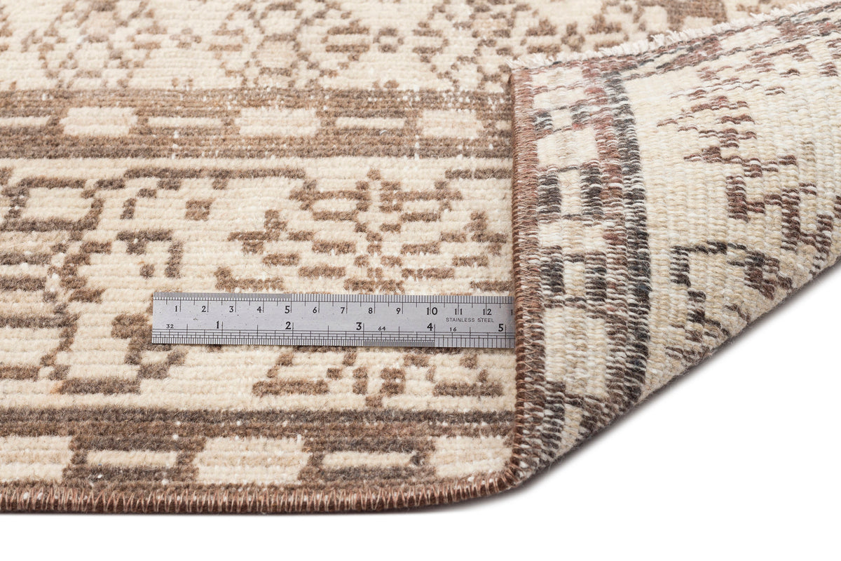 A close-up of the Kidaen - Vintage Medallion Rug (6'0 x 9'6) in beige and brown shows one corner folded to reveal the backing, with a metal ruler on top measuring about 6 inches.