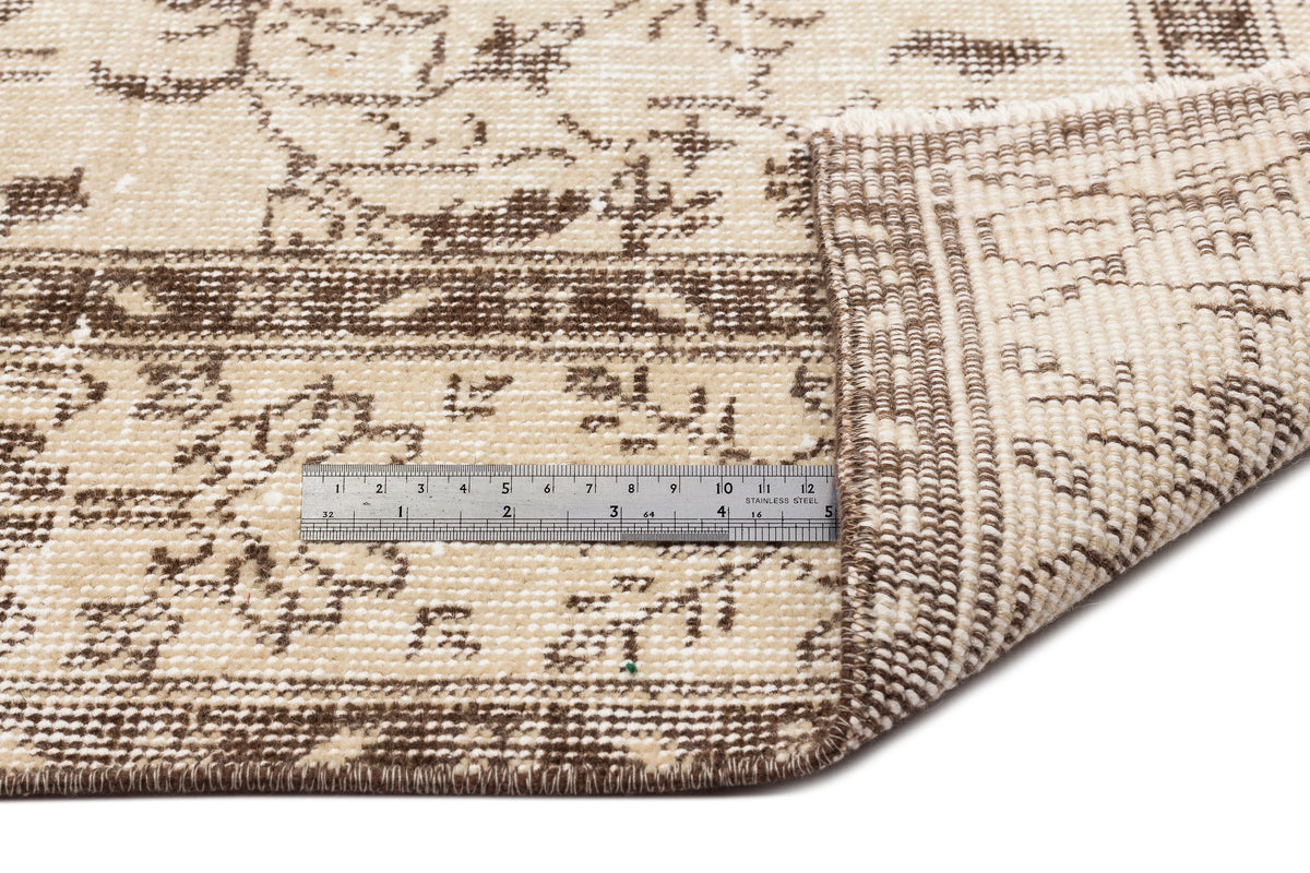 A close-up of the Trabiane - Vintage Floral Rug - 6'1 X 9'11 showcases its beige and brown pattern with one corner folded to reveal the underside. A metal ruler on the rug displays measurements in centimeters.
