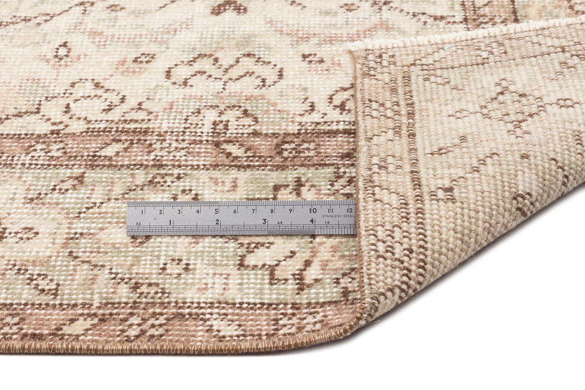 A close-up of the Glela - Vintage Floral Rug (5'0 x 8'11) in beige and brown, with a corner folded to show the underside. A metal ruler beside the fold highlights its thickness and intricate vintage floral design.