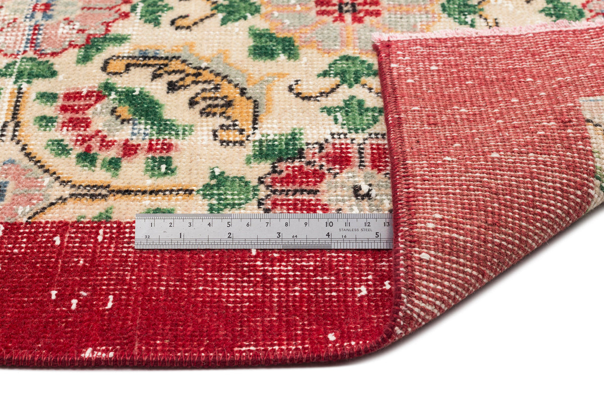Cream rug with red, green, and blue floral pattern, folded corner showing red backing and a metal ruler for scale.