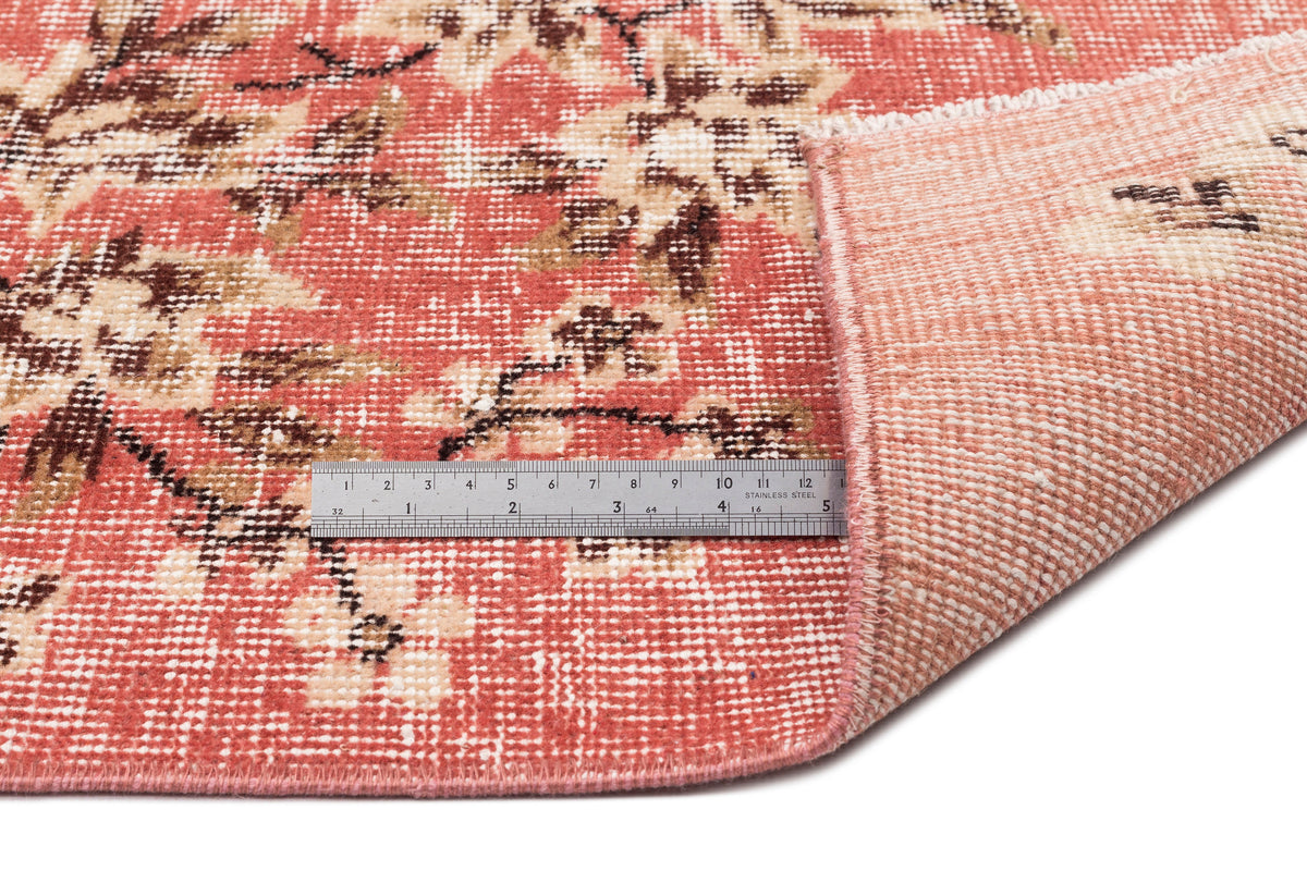 Coral and cream floral patterned rug with a folded corner revealing the woven backing and a metal ruler for scale.
