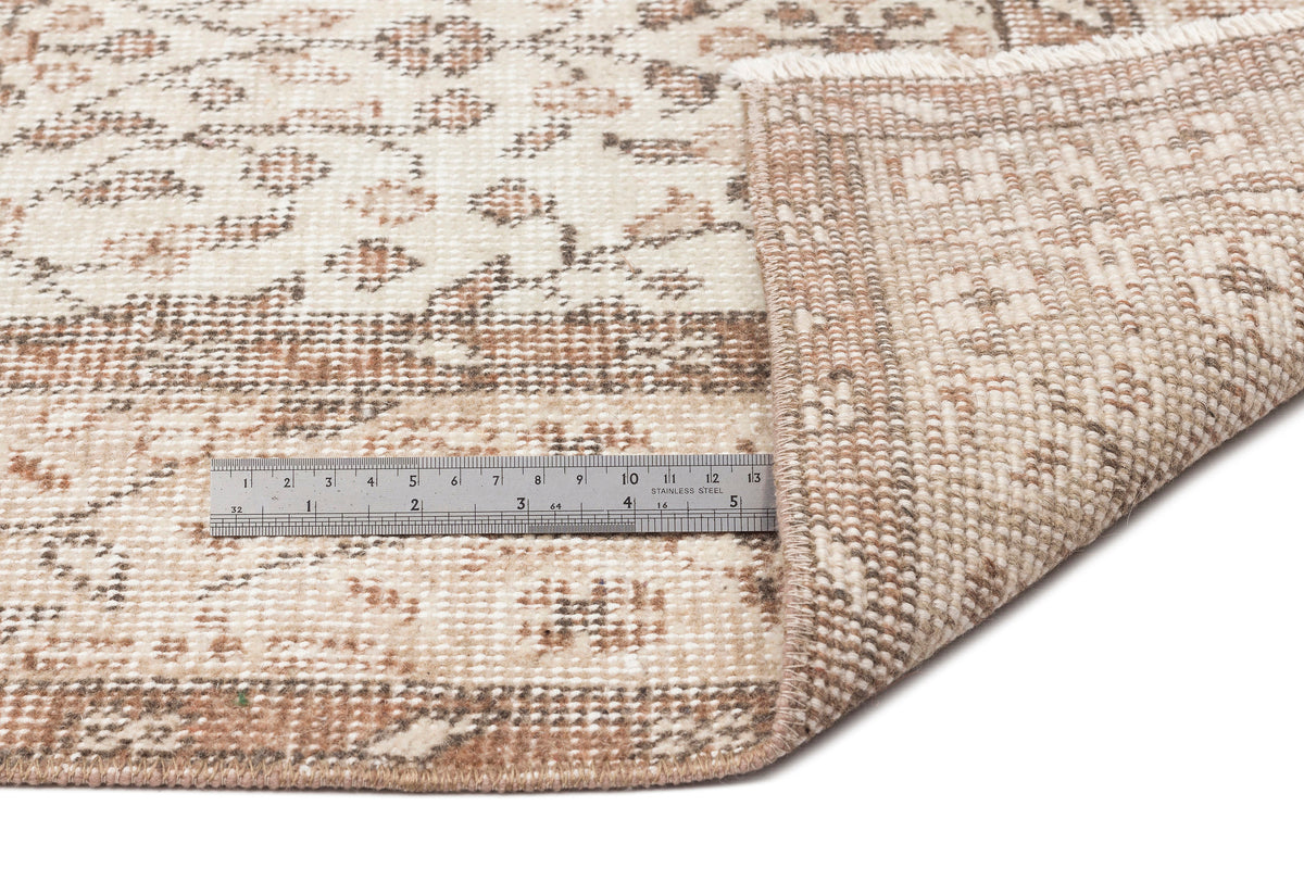 A close-up of the Pavia - Vintage Floral Rug (5'7 x 9'5) in beige and brown, with one corner folded over, next to a metal ruler showing inch and centimeter measurements for scale.