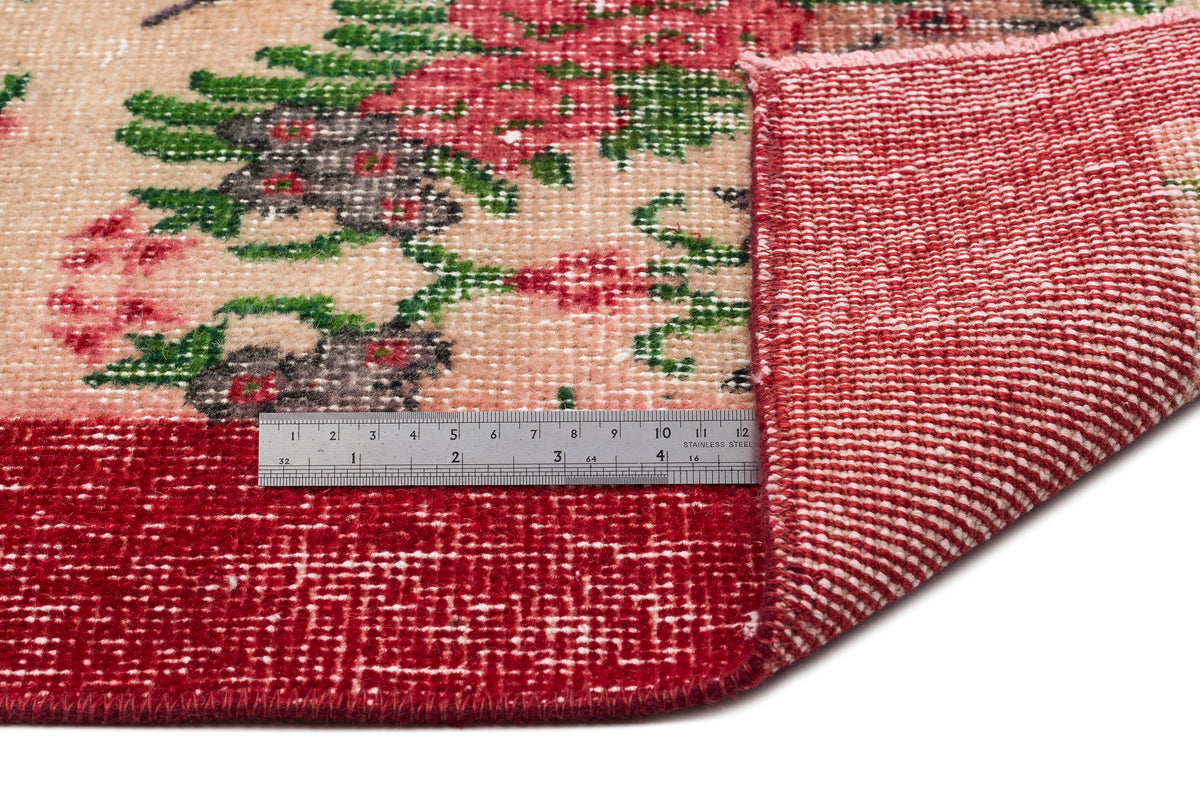 Beige rug with red and green floral pattern, showing a folded corner and a metal ruler for scale on a white background.