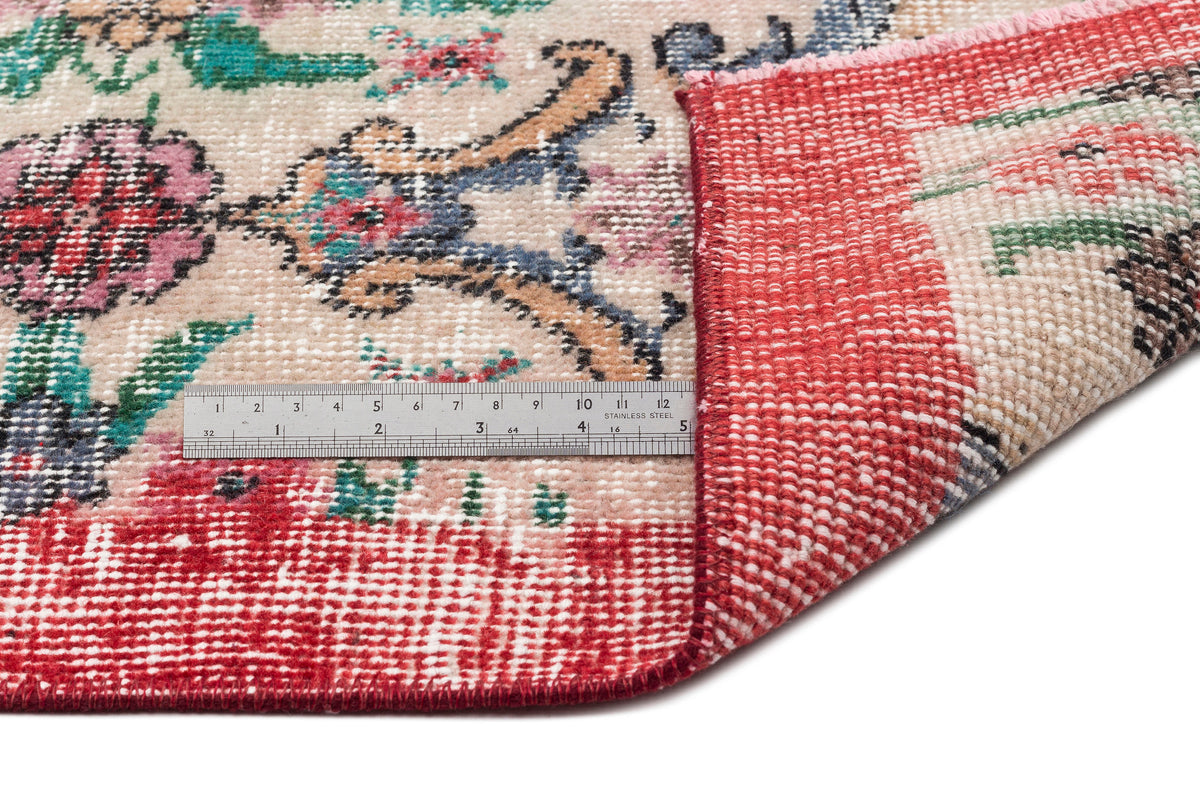 Beige rug with a floral medallion pattern in red, pink, and blue, shown folded to reveal the red woven backing and ruler.
