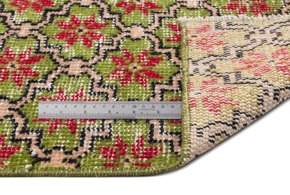 Green, red, and beige rug with a geometric floral pattern, shown with a metal ruler for scale and a folded corner.