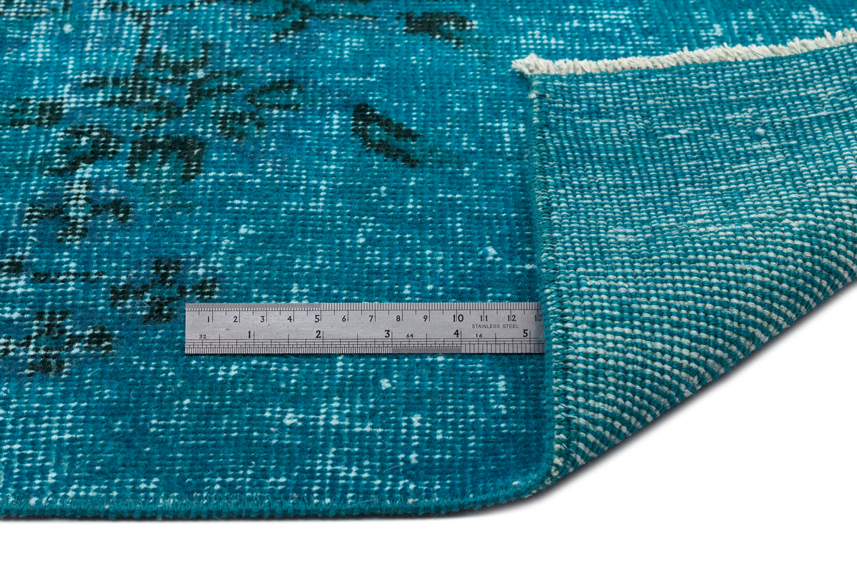 Teal and black distressed rug with a folded corner showing the woven backing and a metal ruler for scale.