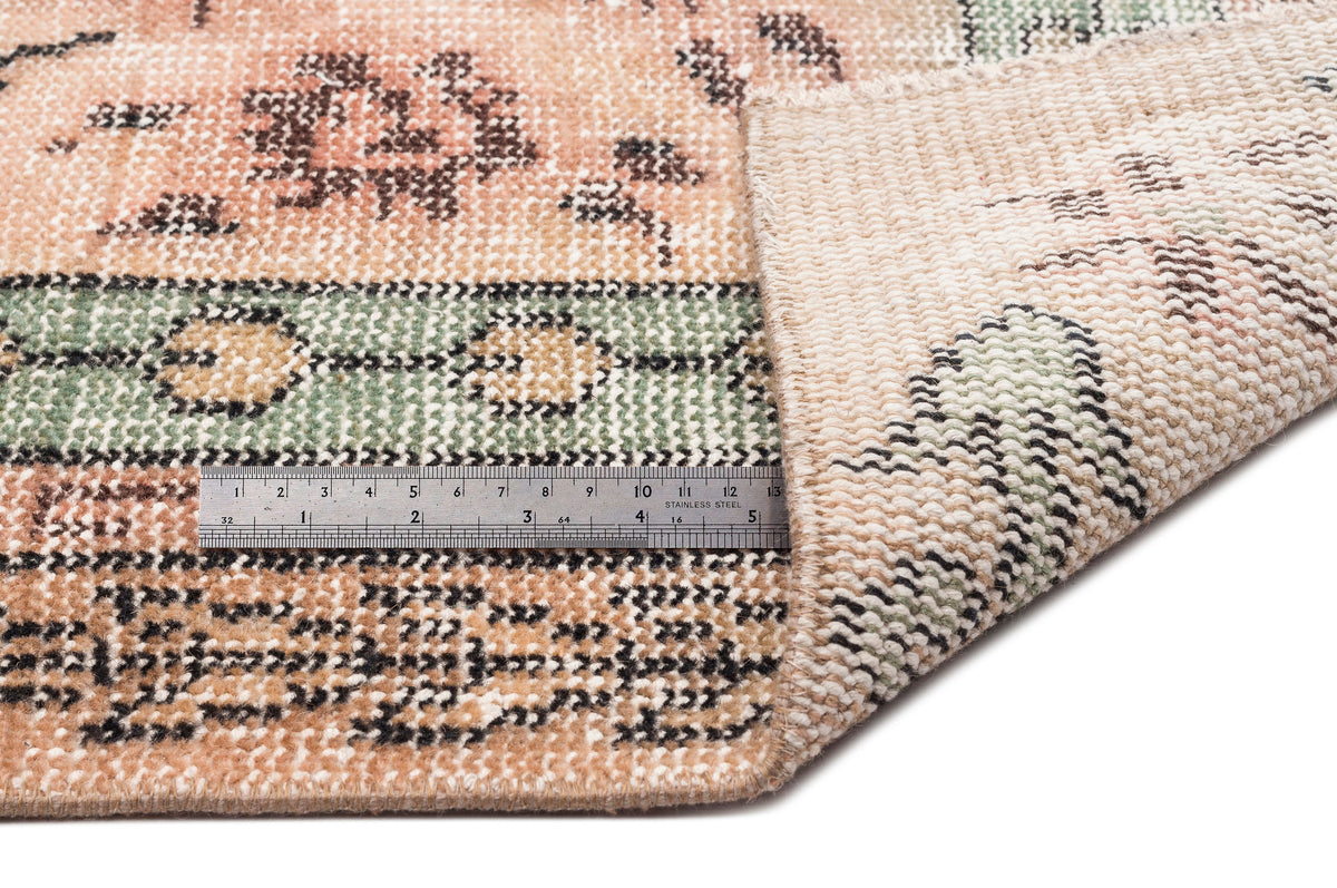 Peach, sage green, and black floral patterned rug with a folded corner and a metal ruler for scale on a white background.
