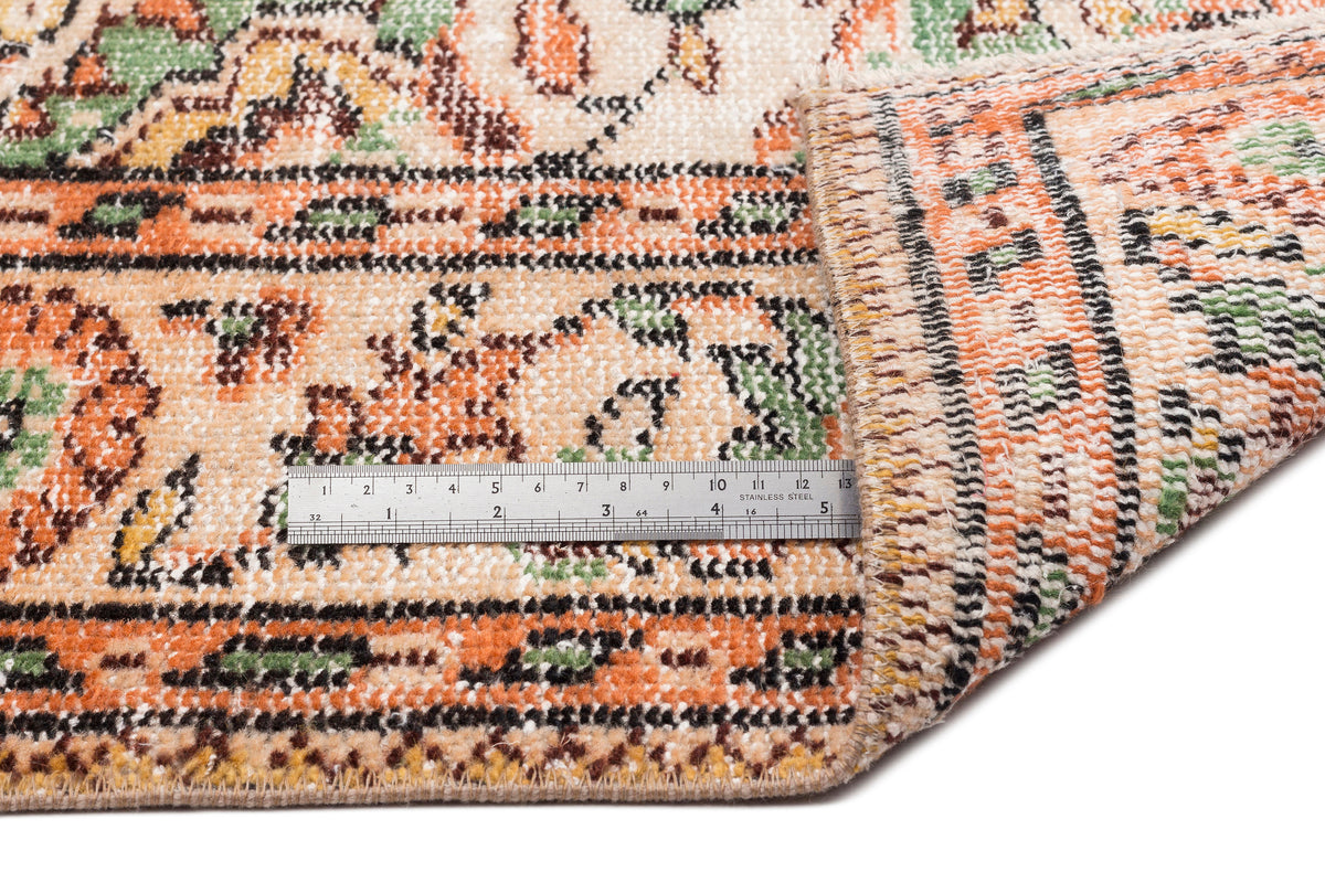 Beige, orange, and green traditional patterned rug with a folded corner and a metal ruler for scale on a white background.