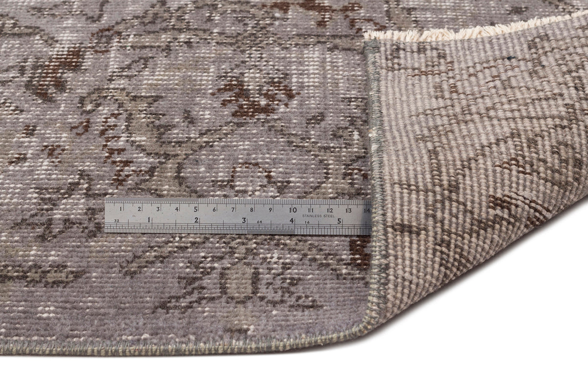 Grey and brown distressed floral rug with a folded corner and a metal ruler for scale on a white background.