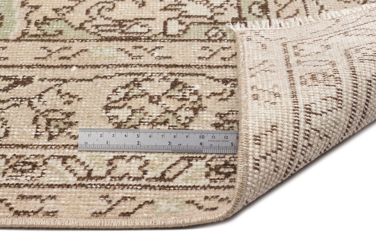 A close-up of the Mamail - Vintage Medallion Rug (6'4 x 9'7) shows its beige patterned surface with a folded corner revealing the back. A metal ruler highlights the rug’s edge, texture, and intricate medallion floral design.